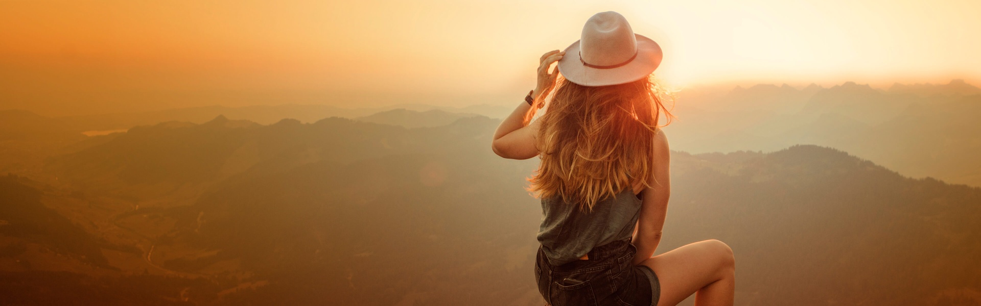 A woman in a hat looking at the sunset on top of a hill.